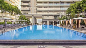 A swimming pool with sun loungers and umbrellas, surrounded by palm trees and a multi-story building with balconies.