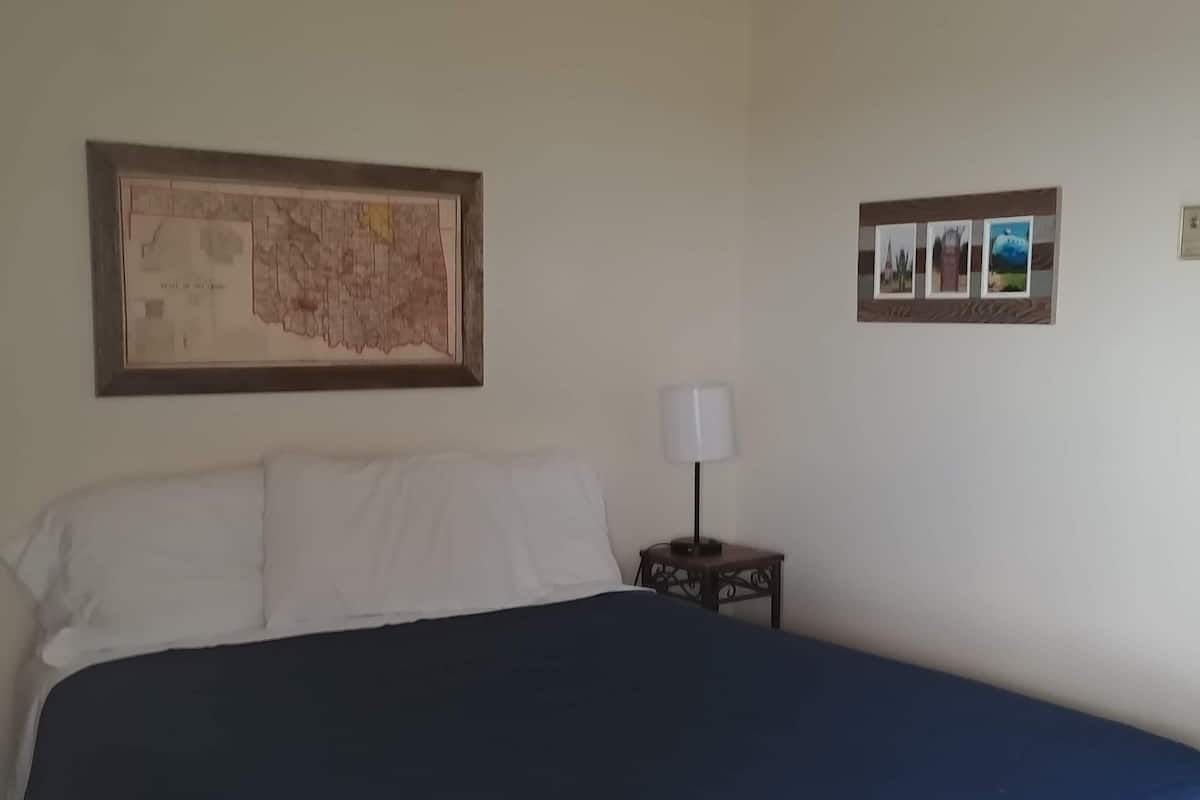 A neatly made bed with a blue bedspread, a framed map on the wall, and a small table with a lamp beside it.