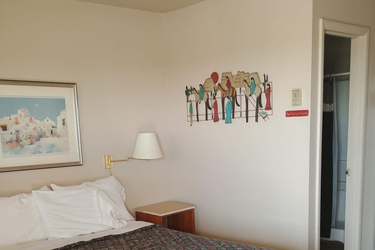 A hotel room with a large bed, a bedside table with a lamp, a framed picture on the wall, and a decorative wall hanging.
