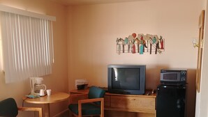 A compact hotel room with a single bed, a small desk with a television and microwave, a chair, a coffee maker, and a wall decoration. - Cactus Inn (McLean)