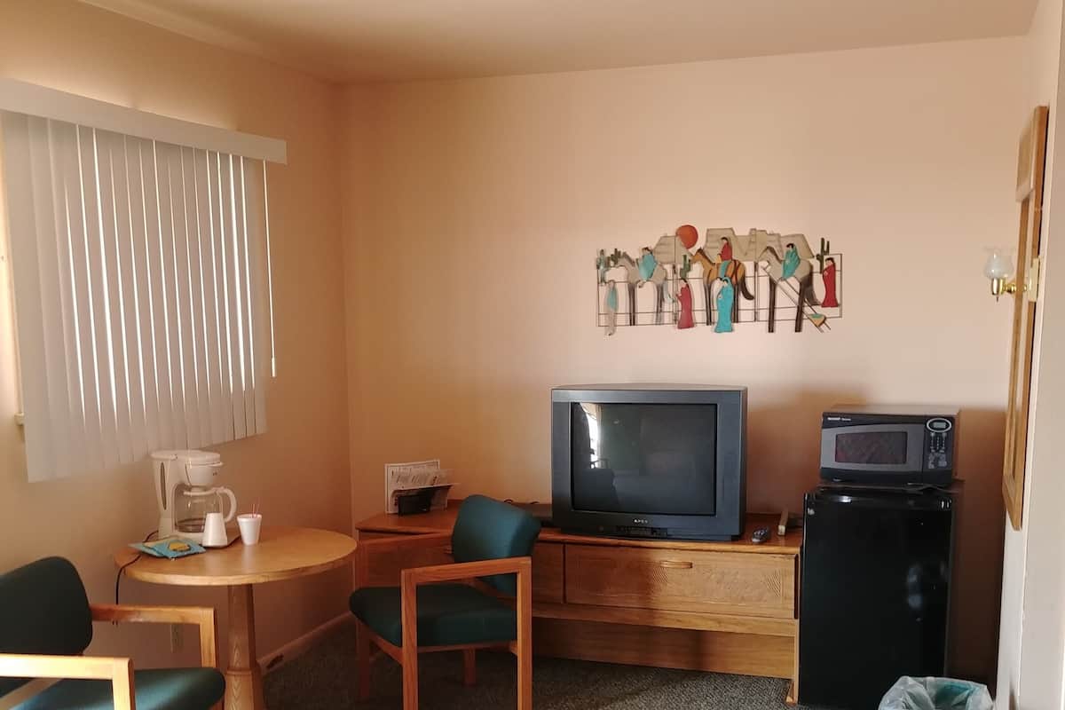 A compact hotel room with a single bed, a small desk with a television and microwave, a chair, a coffee maker, and a wall decoration.