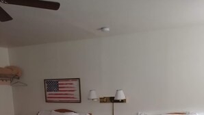 A hotel room with two single beds, a ceiling fan, a nightstand with lamps, and a framed American flag on the wall.