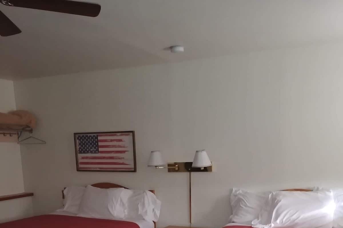 A hotel room with two single beds, a ceiling fan, a nightstand with lamps, and a framed American flag on the wall.