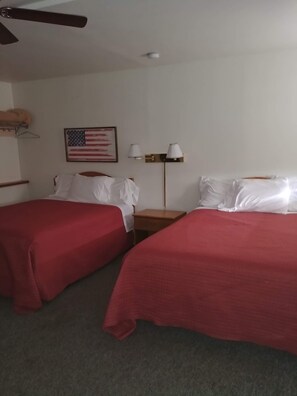 A hotel room with two single beds, a ceiling fan, a nightstand with lamps, and a framed American flag on the wall.