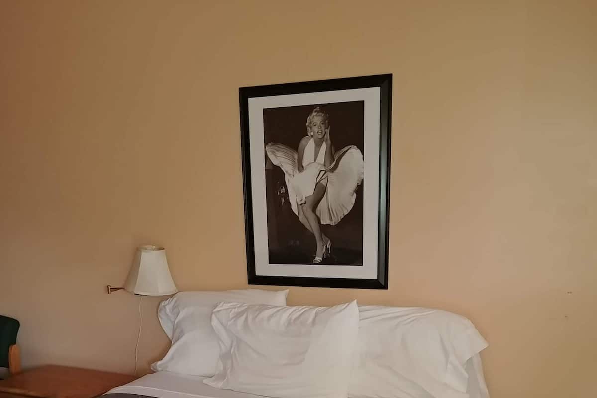 A neatly made bed with a grey quilt, a framed picture on the wall, a bedside lamp, and a wooden nightstand.