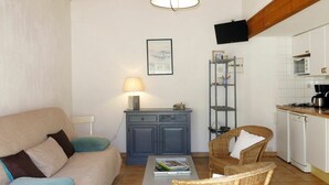 A compact living space with a sofa, a dining table, wicker chairs, a small kitchen area, and a wall-mounted TV. - Cosy apartment for 4 people with WIFI, TV and terrace (La Grainetière)