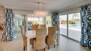 Uma sala de jantar com cadeiras de vime e uma mesa branca, janelas amplas e vista para uma piscina.