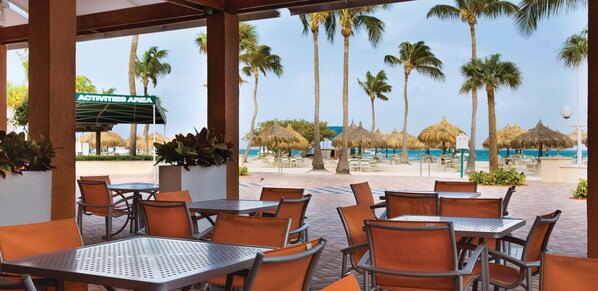 A covered outdoor seating area with tables and chairs, overlooking a beach with palm trees and a sign for activities.