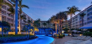 A resort pool area with a curved pool, palm trees, and a building with multiple balconies.