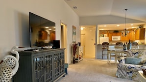 A living room with a TV on a cabinet, a sofa, and a dining area with chairs and a table. - Perfect Location 2/2 Villa in Creekside Landing walking distance to Lake Sumter (The Villages)