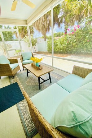 Relax in a serene patio setting with a comfortable seating area, featuring a sofa and wicker chairs, and enjoy the view of a lush garden.