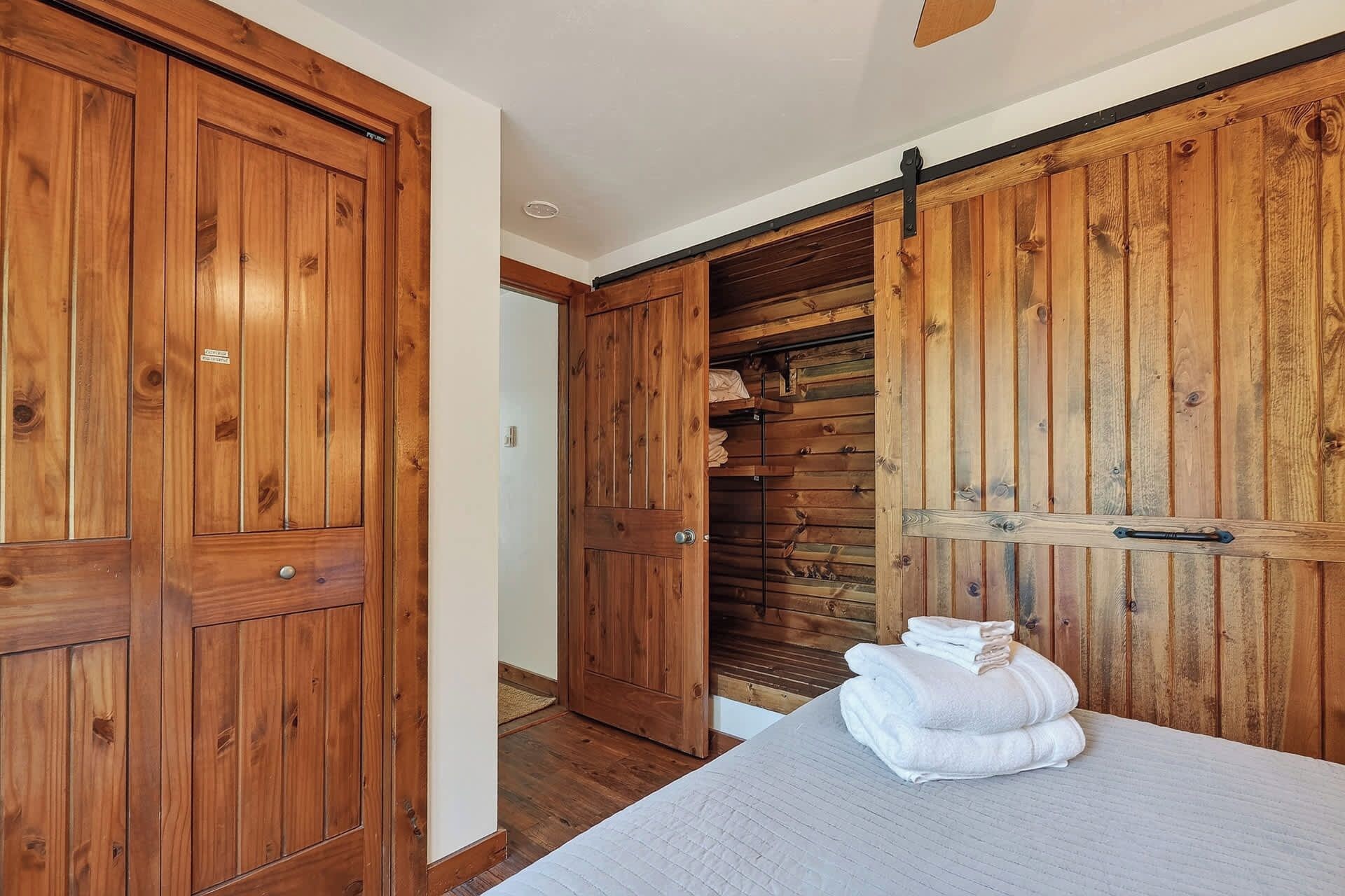 Une pièce avec des armoires en bois, un lit recouvert d’une couette blanche et une porte en bois donnant sur un couloir.