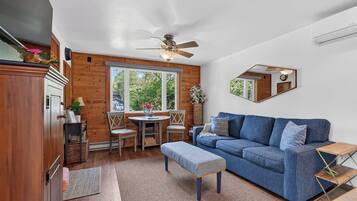 Une pièce avec un canapé bleu, des meubles en bois, une table à manger avec des chaises, un ventilateur de plafond et une grande fenêtre.