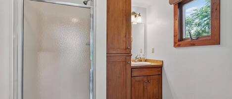 A bathroom with a shower, wooden cabinets, a sink, and a window with a view.
