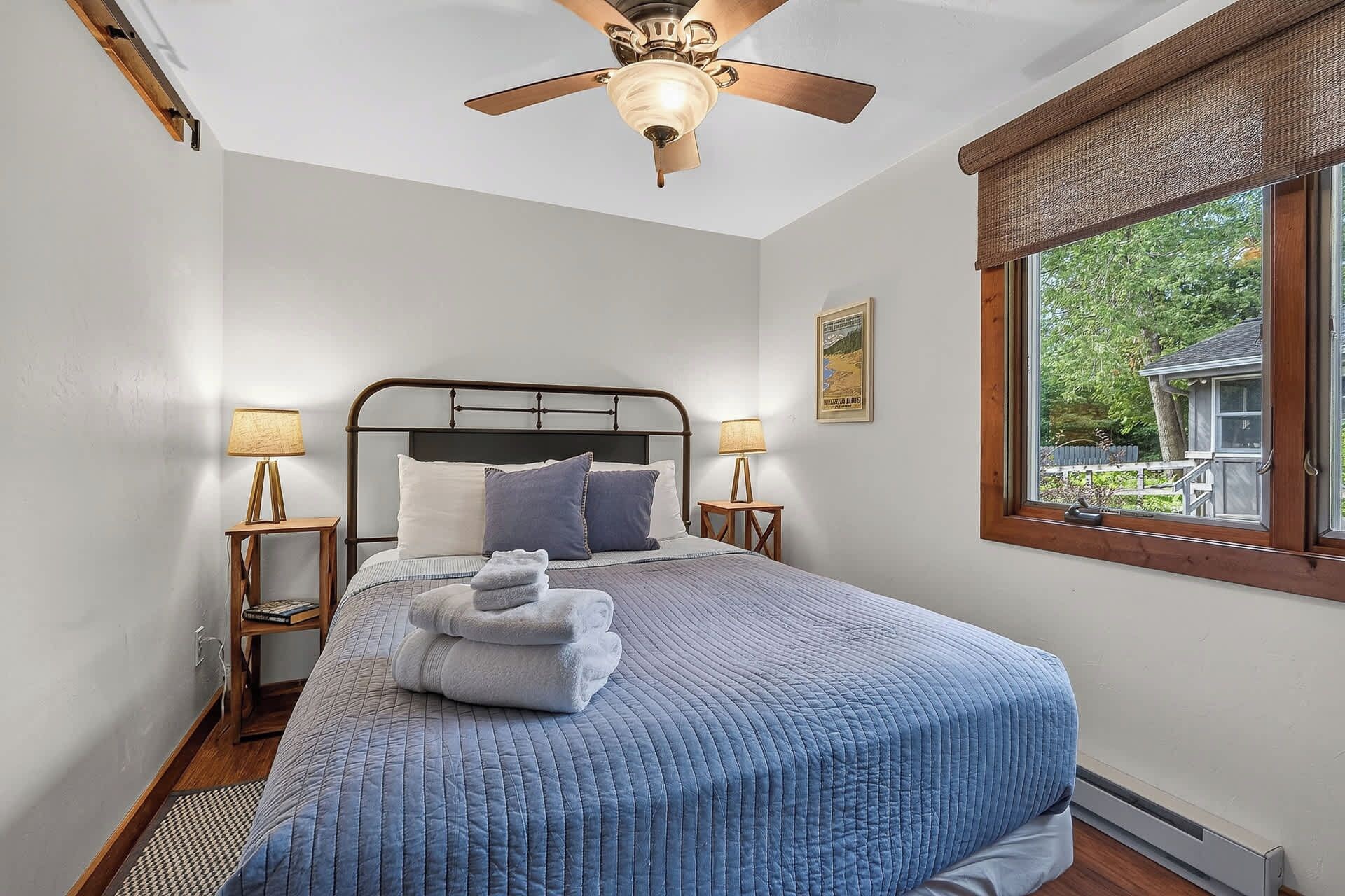 A bedroom with a bed, two bedside lamps, a ceiling fan, a window with a view, and a framed picture on the wall.