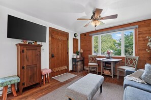 A cozy room with a wooden floor, a ceiling fan, a television, a wooden wardrobe, a bench, a table with chairs, and a window with a view of trees. - Cabin in Downtown Ephraim-Perfect for couples! (Ephraim)