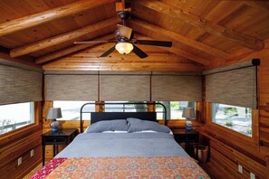 Une chambre de style cabane en bois, avec un lit, deux tables de chevet, un ventilateur de plafond et de grandes fenĂȘtres Ă©quipĂ©es de stores.