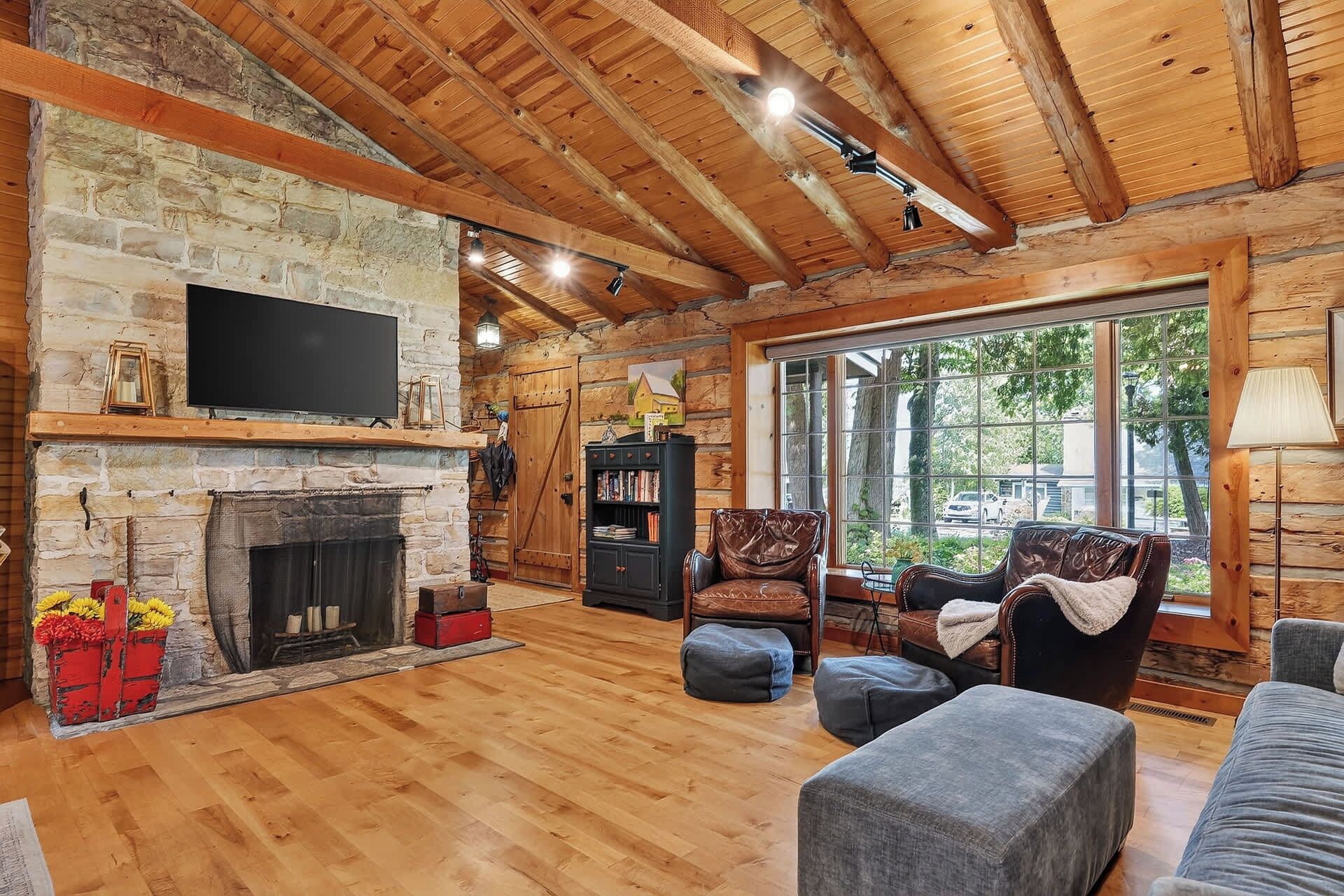 A rustic cabin interior with a stone fireplace, wooden beams, a large window, and a cozy seating area.