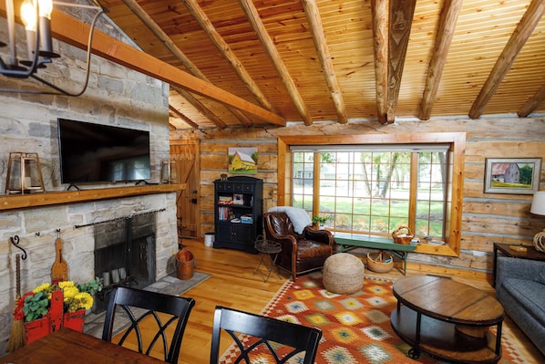 A log cabin interior with a stone fireplace, wooden ceiling beams, a television, and a large window. - Remodeled Log Cabin | Dog-Friendly Near Wilson's (Ephraim)
