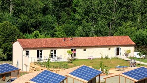 Ein Gebäude mit rotem Ziegeldach, umgeben von Solaranlagen, vor einer Kulisse aus dichtem Grünwerk.