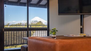 A room with a large window offering a view of a mountain, a flat-screen TV mounted on the wall, and a small table with a plant and a sign. - IzuRicca (Izunokuni)