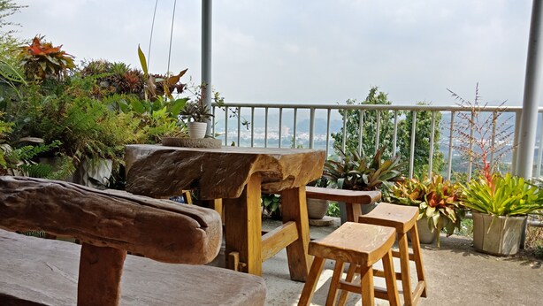 Un área de estar al aire libre con bancos y una mesa, rodeada de plantas en macetas y vista a un paisaje urbano.