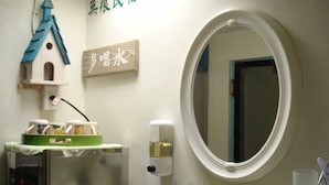 Un baño con un espejo redondo, un lavamanos pequeño, un refrigerador, una decoración con forma de casita para pájaros y una planta en maceta.