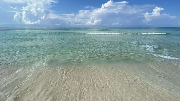 A beach with clear water, sandy shore, and a blue sky with clouds.