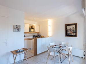Une cuisine compacte avec des meubles blancs, un four à micro-ondes et une machine à café. On y trouve une table à manger avec quatre chaises et une petite table d’appoint avec des tasses.