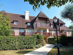 Une grande maison à plusieurs étages, dotée d’un balcon imposant au deuxième étage, entourée de haies bien entretenues et baignée par un ciel dégagé.