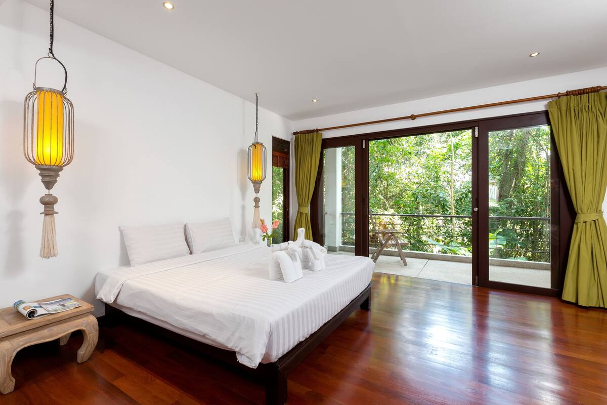 A spacious bedroom with a large bed, wooden flooring, and a balcony with green curtains.