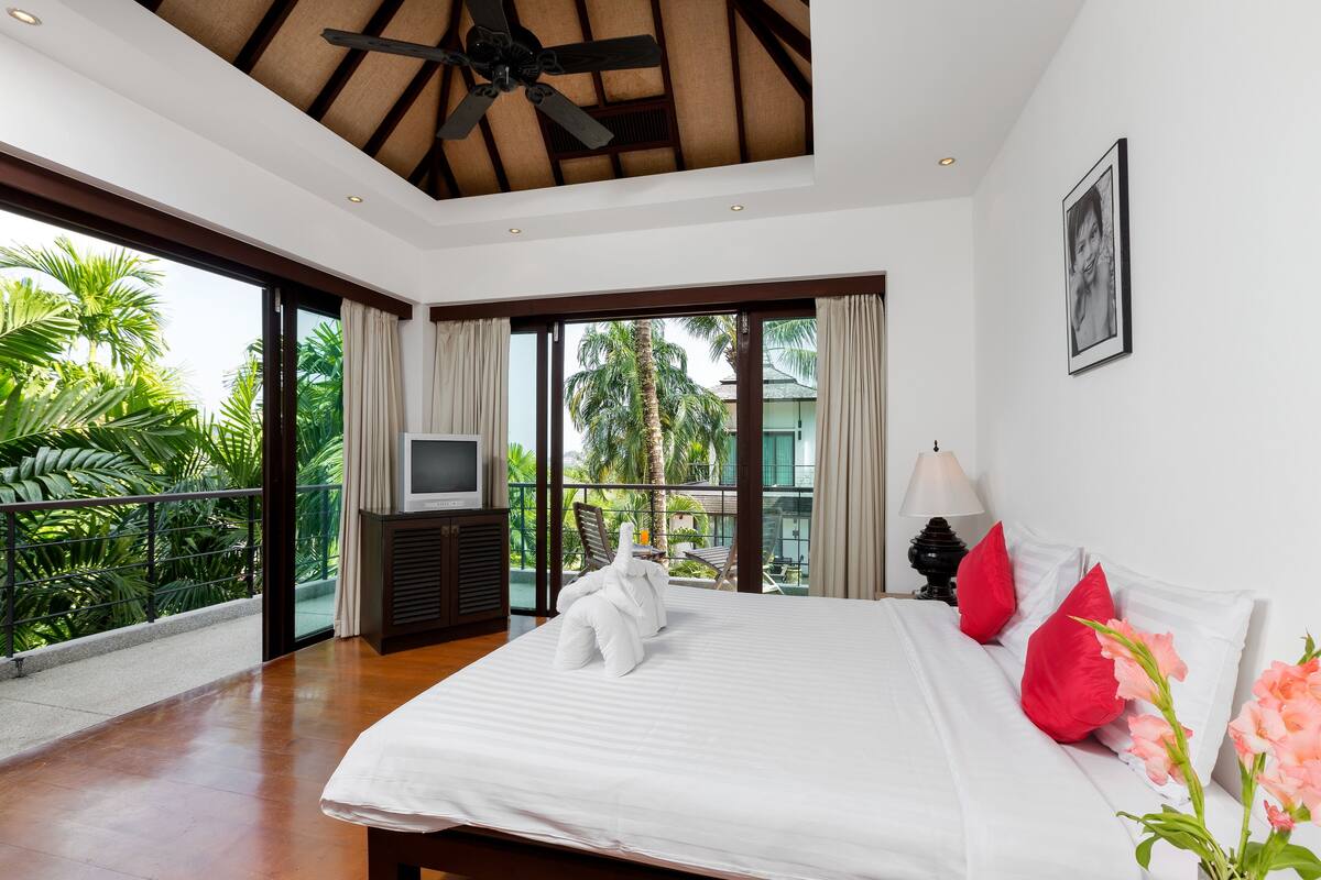 A spacious bedroom with a large bed, a ceiling fan, and a balcony with a view of palm trees.