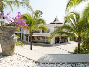Un edificio moderno con un amplio ventanal de vidrio, rodeado de palmeras y una jardinera de piedra con flores moradas.