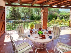 Une terrasse extérieure couverte, dotée d’une table ronde et de six chaises, entourée d’un mur en pierre et d’une rambarde en métal.