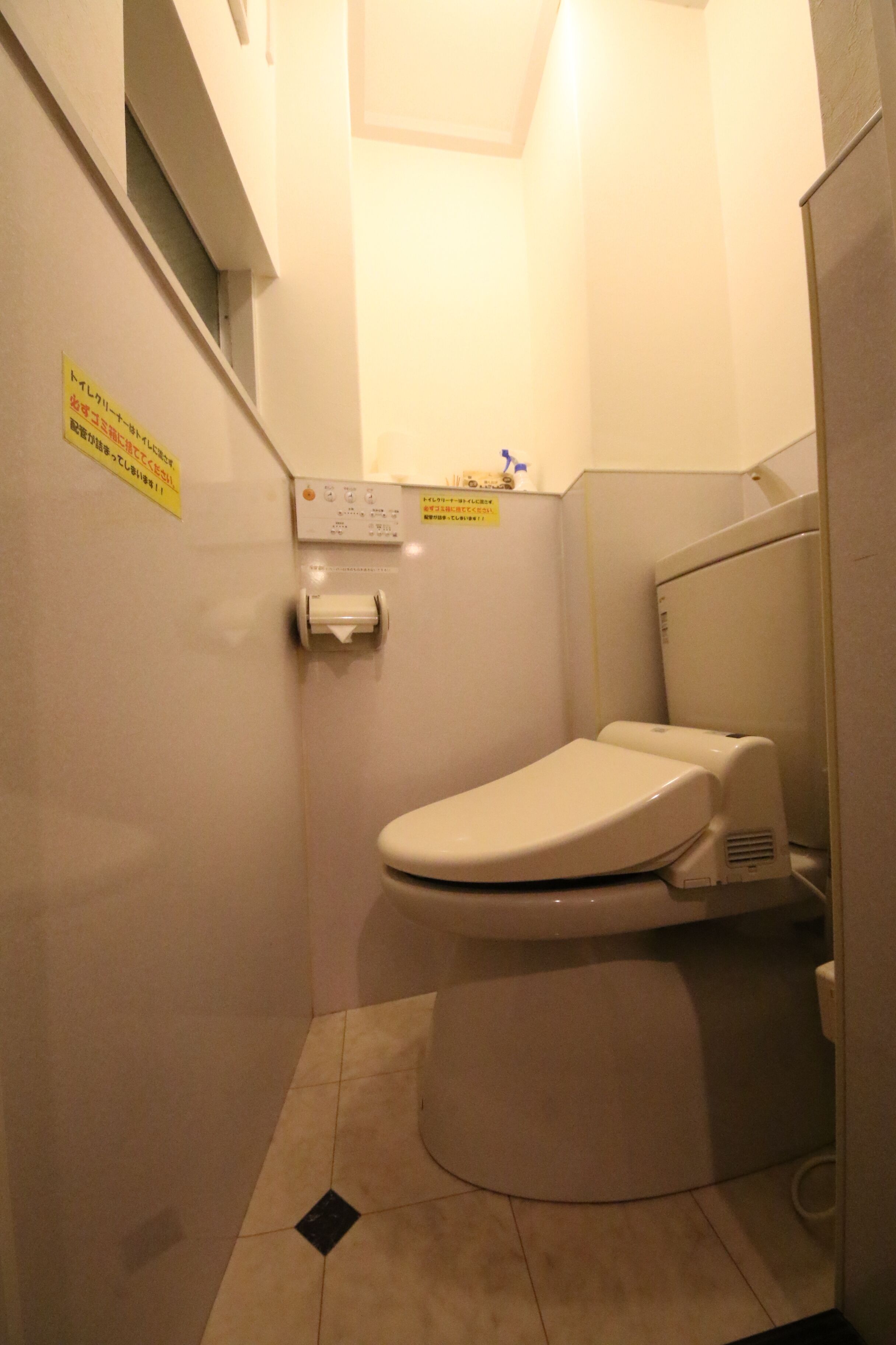 Casa de banho pequena e compacta com sanita, secador de mãos e dispensador de papel. Há uma janela sobre a sanita e um sinal na parede.