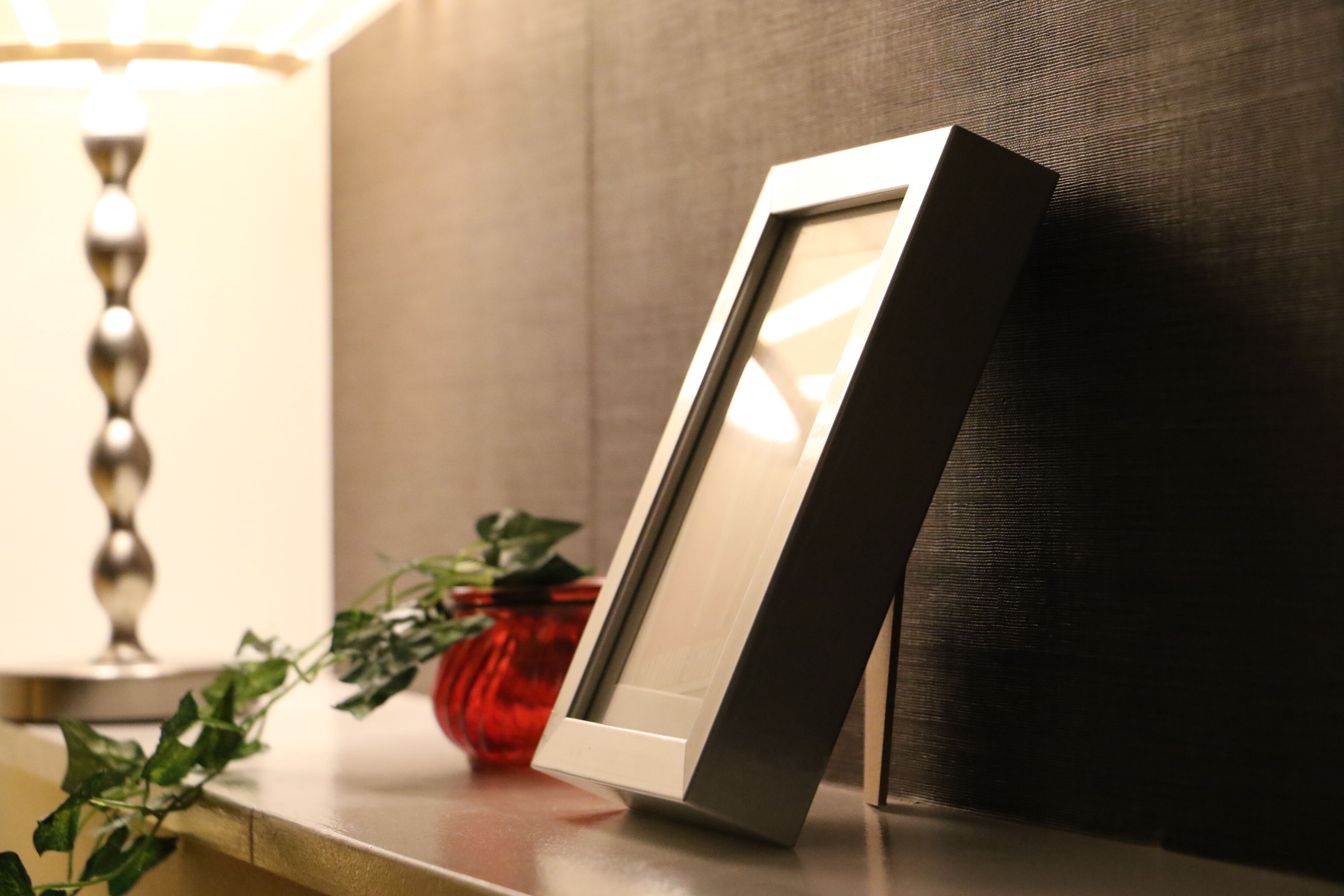 Un tableau encadré appuyé contre un mur, un vase rouge avec une plante, et une lampe en arrière-plan.