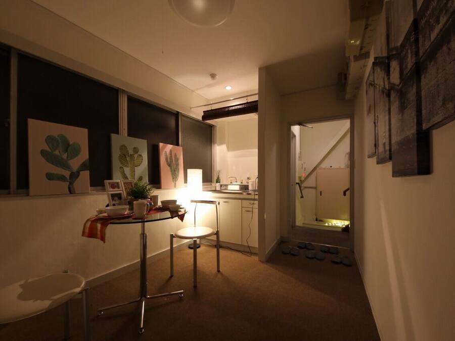 Un pasillo estrecho con una kitchenette a la derecha, un comedor con mesa y sillas, y una escalera que sube a otro piso.