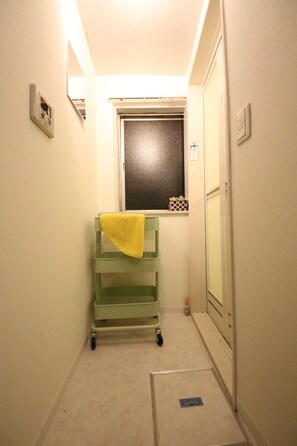 Un pasillo estrecho con un carrito verde, una ventana con persiana de cuadros y una puerta con interruptor de luz.