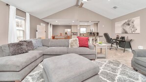 Un salon spacieux avec un canapé d’angle gris, une petite table et un ventilateur de plafond.