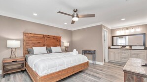 Une chambre à coucher avec un lit en bois, des tables de chevet, un ventilateur de plafond et une salle de bain équipée d’un meuble-lavabo et d’un miroir.