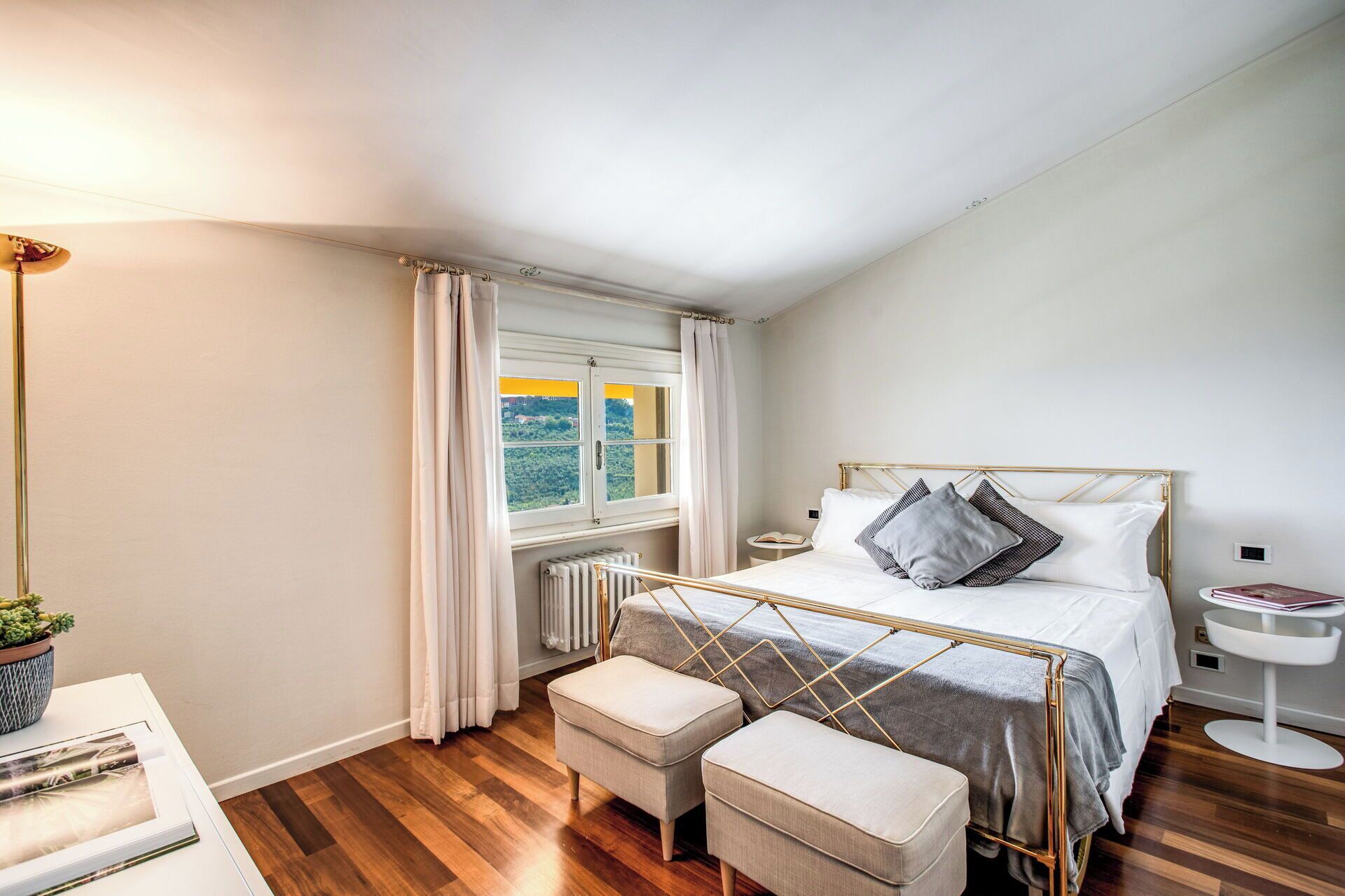 Ein Schlafzimmer mit einem Bett, zwei Nachttischen, einem Heizkörper, einem Fenster mit Vorhängen und einem kleinen Tisch mit einer Pflanze.