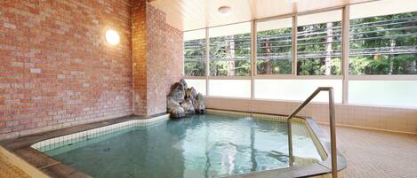 An indoor swimming pool with a brick wall and tiled floor, large windows with a view of trees, and a metal ladder for access.