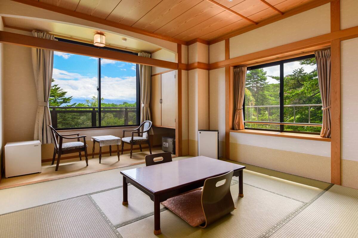 A traditional Japanese-style room with tatami flooring, a low table, and chairs. Large windows offer a view of greenery.