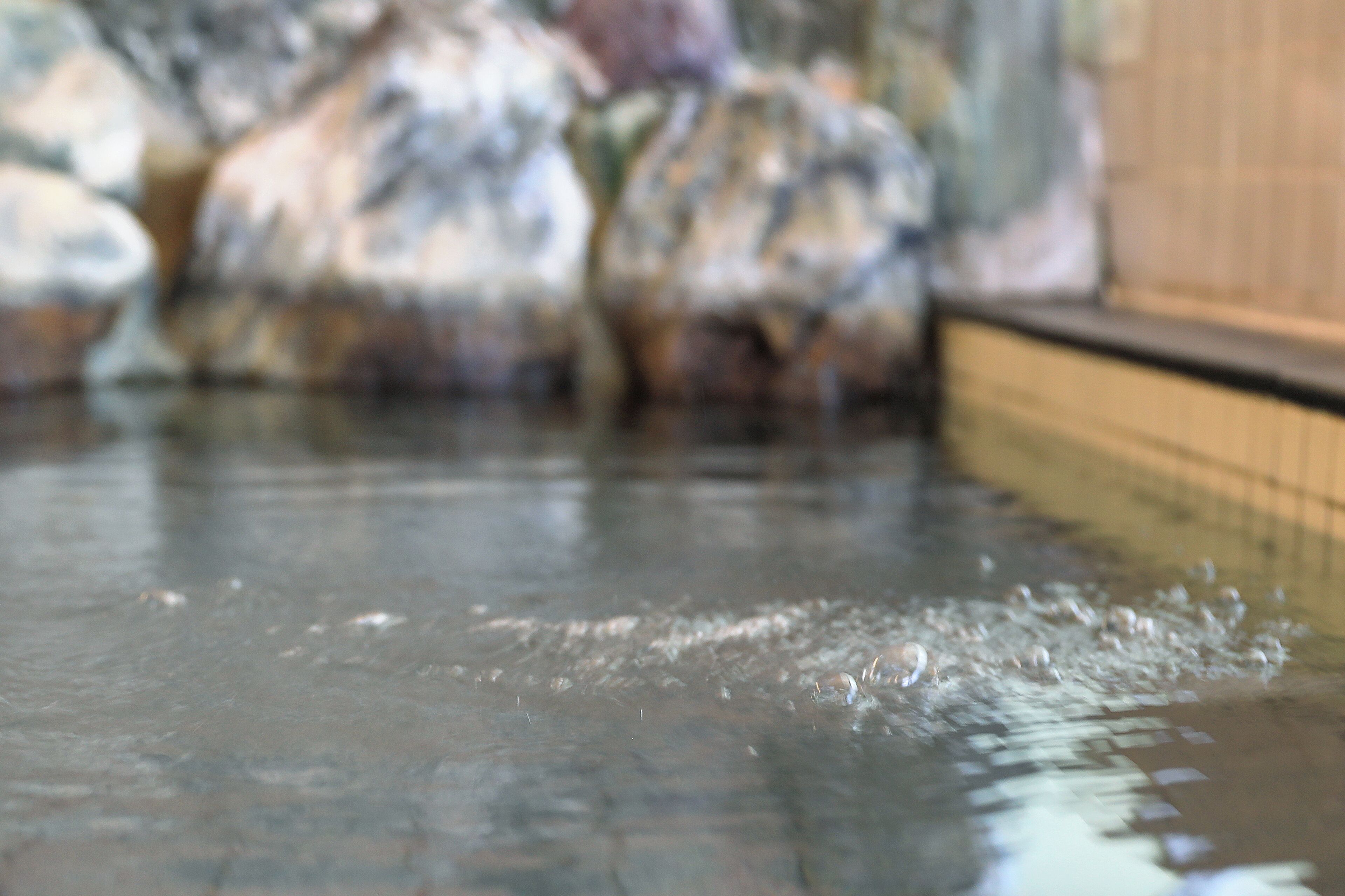 Eine Nahaufnahme einer Wasseroberfläche mit Blasen, vermutlich aus einer Thermalquelle oder einem Pool, mit unscharfem Hintergrund aus Felsen und einer Holzkonstruktion.