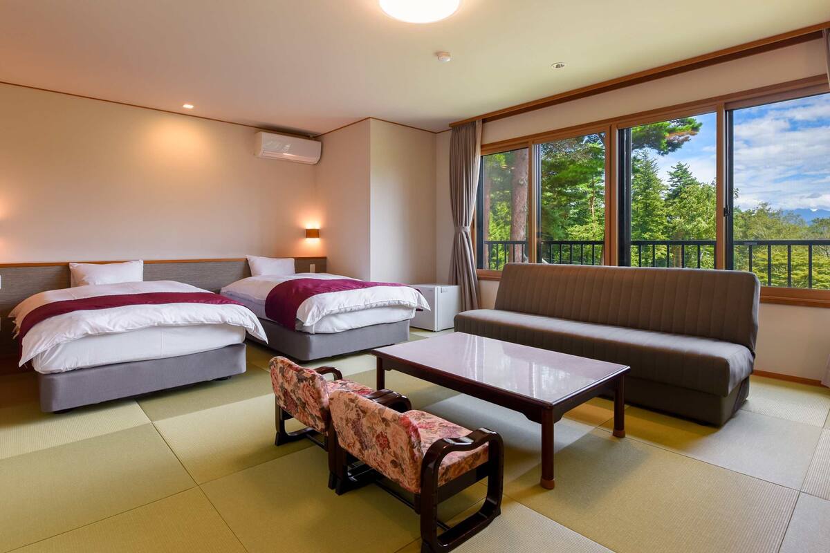 A hotel room with two beds, a sofa, a small table, tatami mats, and a large window with a view of trees.