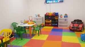 Une salle de jeux pour enfants avec un sol coloré, des chaises vertes, une table et divers jouets.