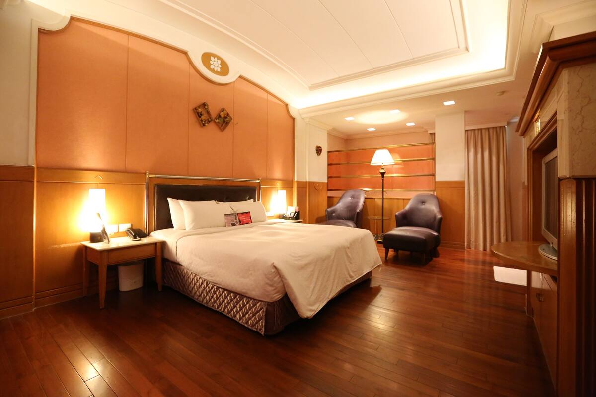 A hotel room with a large bed, two armchairs, a desk with a lamp, and a television.
