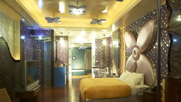 Ein modernes Hotelzimmer mit einem großen Bett, einem Fernseher, einem Deckenventilator und einem im Hintergrund sichtbaren Badezimmer.