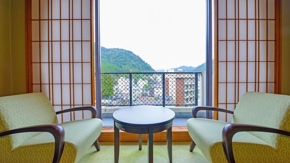 A room with two chairs and a table, a view of a balcony with a fence, and a mountain in the background.