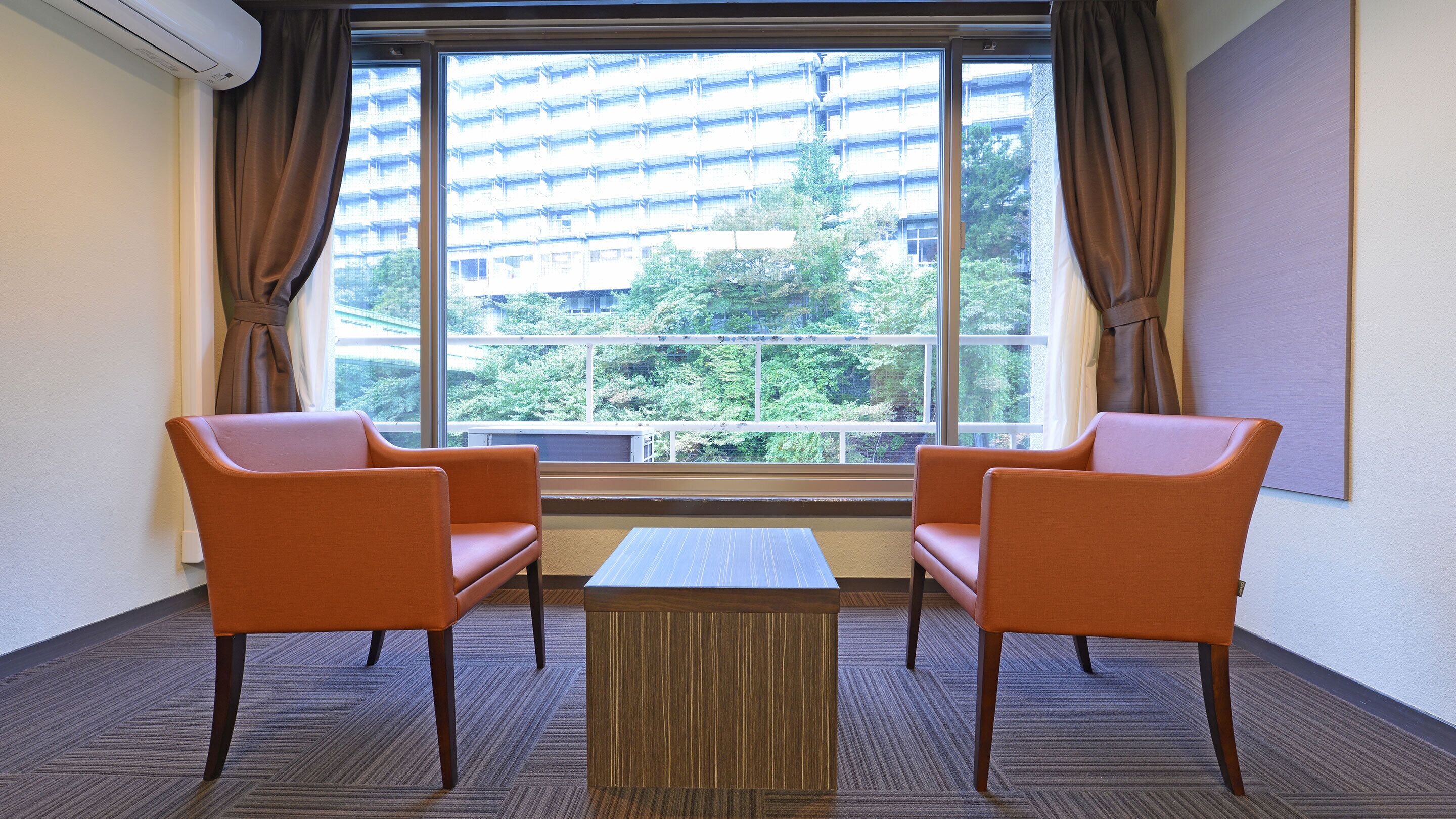 Ein Hotelzimmer mit zwei orangefarbenen Sesseln, einem kleinen Holztisch und einem großen Fenster mit Vorhängen.